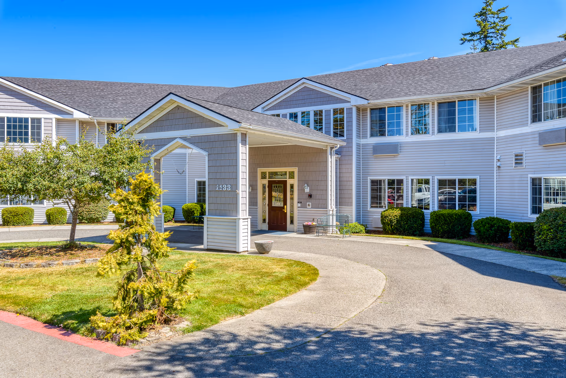 Front entrance of a two-story assisted living building with a covered porte-cochère, windows, and a landscaped circular driveway.