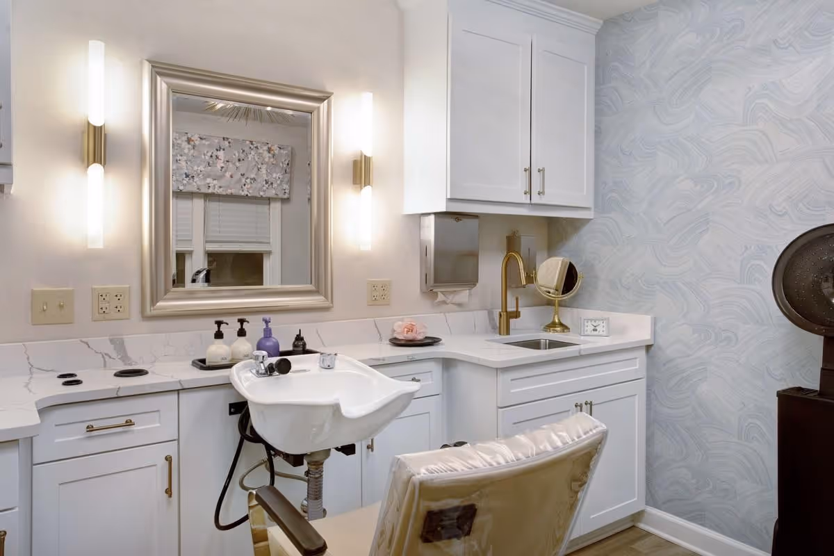 Salon-style hair-wash station with a white sink and chair in front of a mirror, white cabinetry, countertop and gold fixtures.
