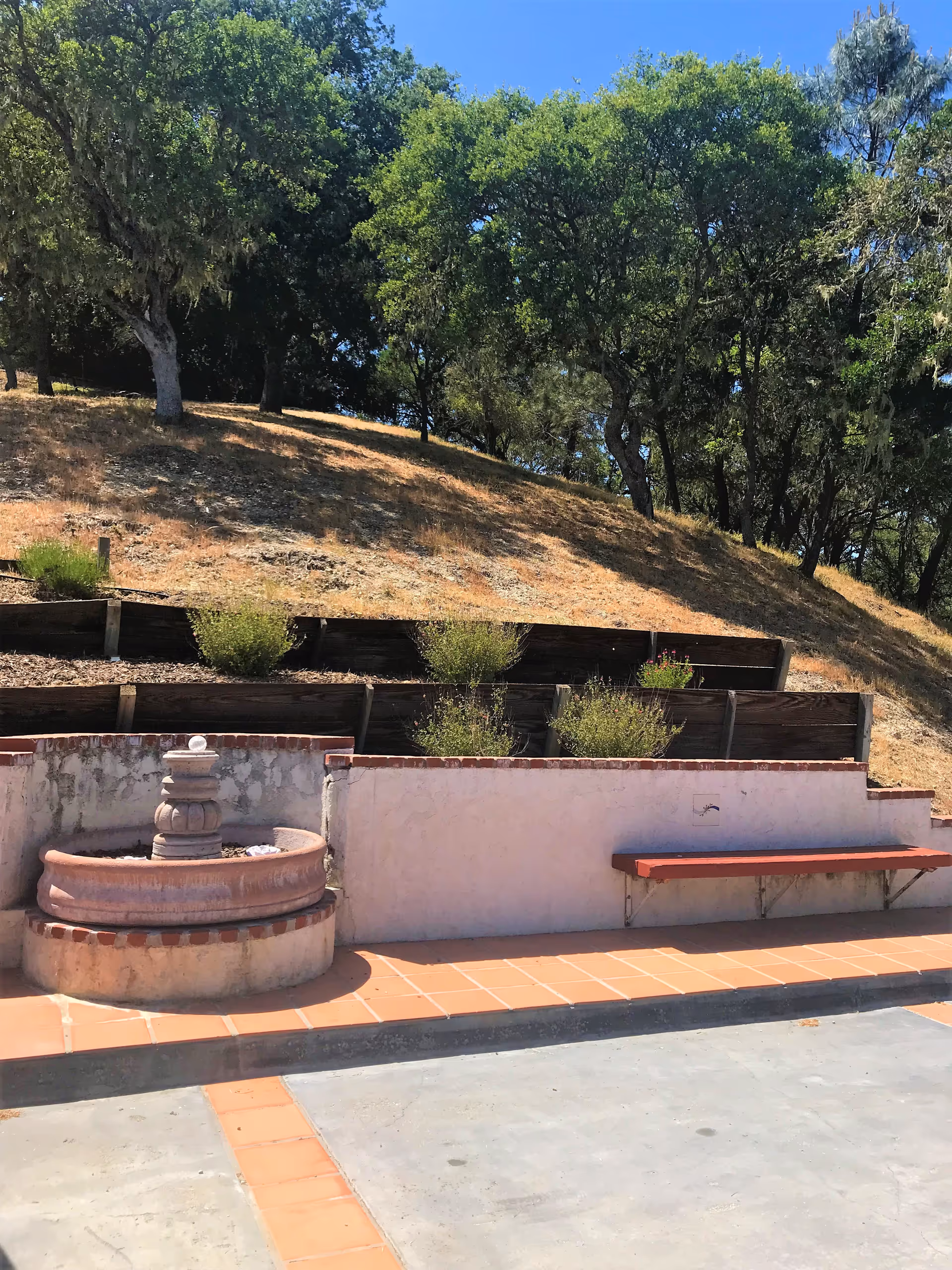 Outdoor area with a small tiered fountain, a long bench attached to a white stucco wall with a red top, terracotta tile flooring, and a hillside with trees and shrubs in the background under a clear blue sky.