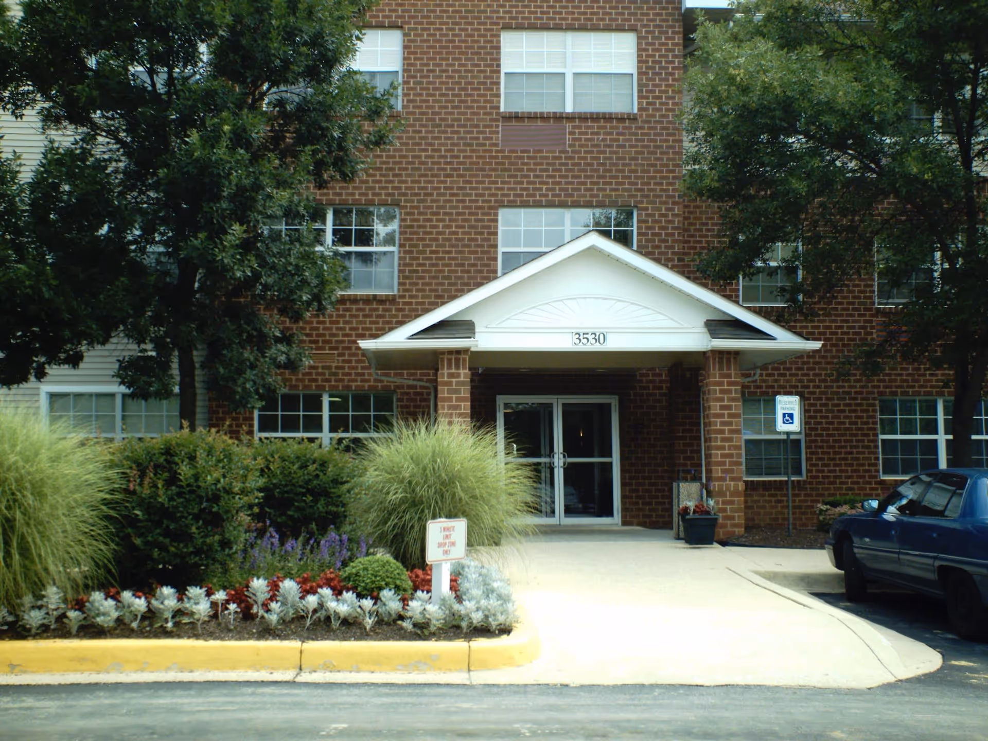 Brick senior living building entrance with a covered portico labeled 3530, landscaping, and a parked car.