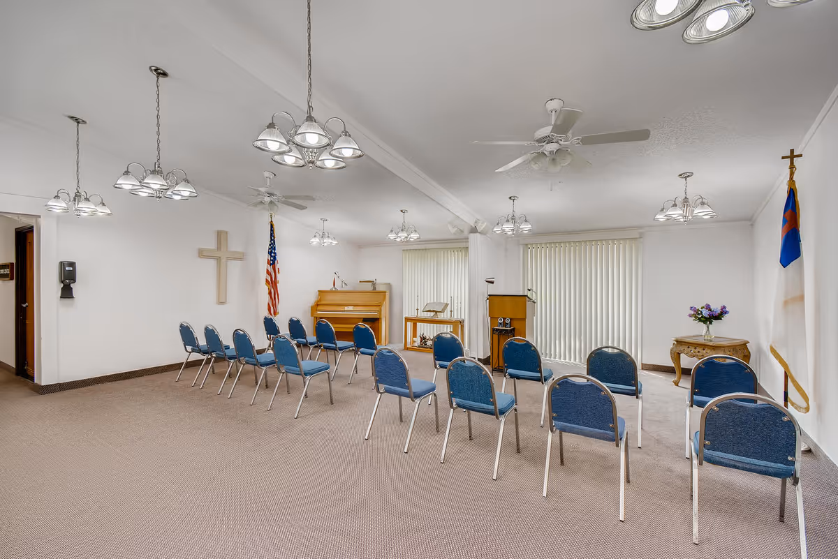 A small chapel or meeting room with rows of blue chairs arranged facing a wooden piano, a lectern, and two flags on either side of the room. The room has beige carpet, white walls, ceiling fans, and multiple hanging light fixtures. There is a wooden cross on one wall and a small table with a vase of flowers on the other side.
