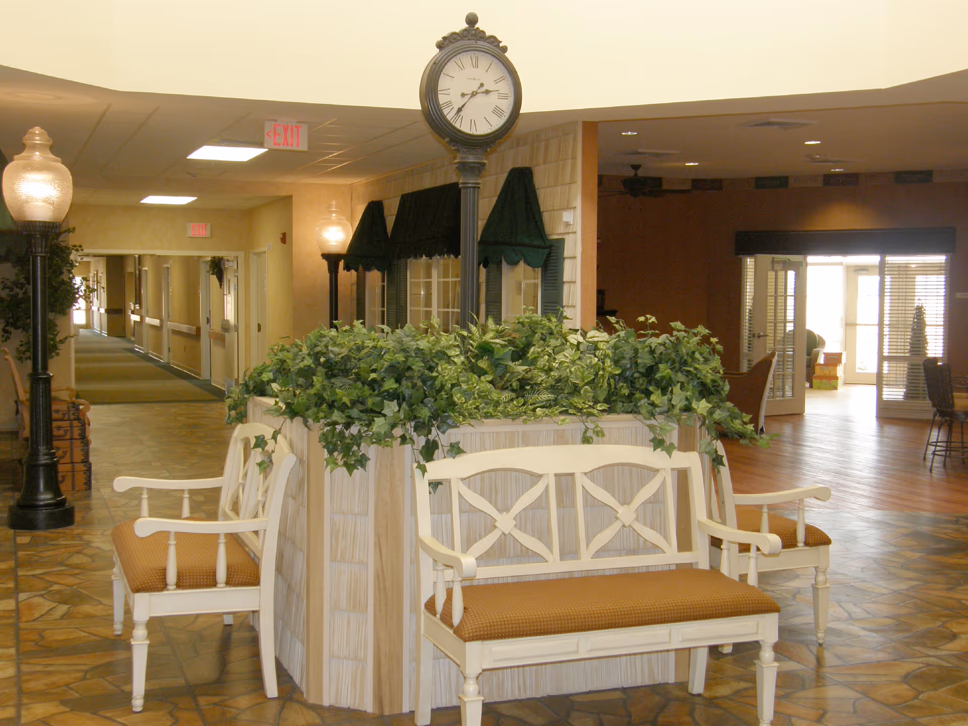 Interior view of a senior living facility hallway with a central seating area featuring white wooden benches with brown cushions surrounding a planter filled with green leafy plants. A decorative black street clock stands in the middle of the planter. The hallway has tiled flooring, beige walls, and ceiling lights. There are two black lamp posts with glass globes on either side of the planter. In the background, there is an open area with wooden flooring and glass doors letting in natural light.