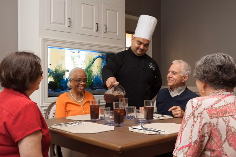 A chef in a black uniform pours a beverage into a glass at a dining table where four elderly people are seated, smiling and engaging with each other. The setting is indoors with a large fish tank built into the wall behind them.