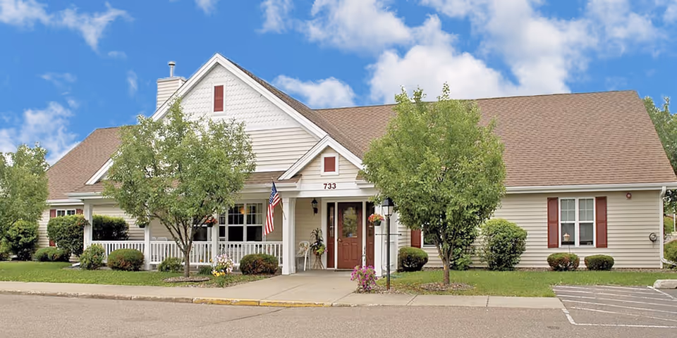 Exterior front view of a single-story senior living facility building with beige siding, a brown shingled roof, and red shutters. The entrance has a covered porch with white railings, an American flag, and some landscaping including trees and bushes. The building number 733 is visible above the door.