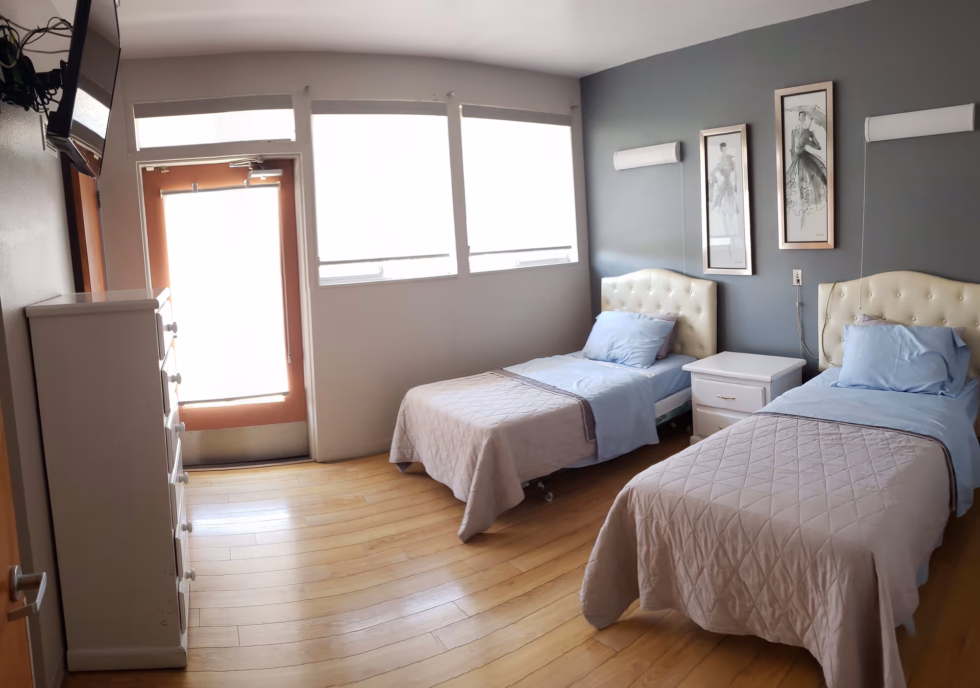 A bright, clean bedroom with two single beds featuring tufted headboards, light blue bedding, and beige quilts. Between the beds is a white nightstand with two drawers. The room has light wooden flooring, a white chest of drawers on the left, and a door with windows above it letting in natural light. Two framed artworks hang on the gray accent wall behind the beds.