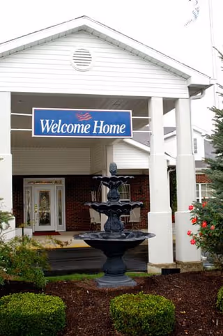 Entrance of a senior living facility with a white covered porch, a black tiered fountain in front, and a blue sign hanging that reads 'Welcome Home'. There are bushes and flowers around the entrance area.