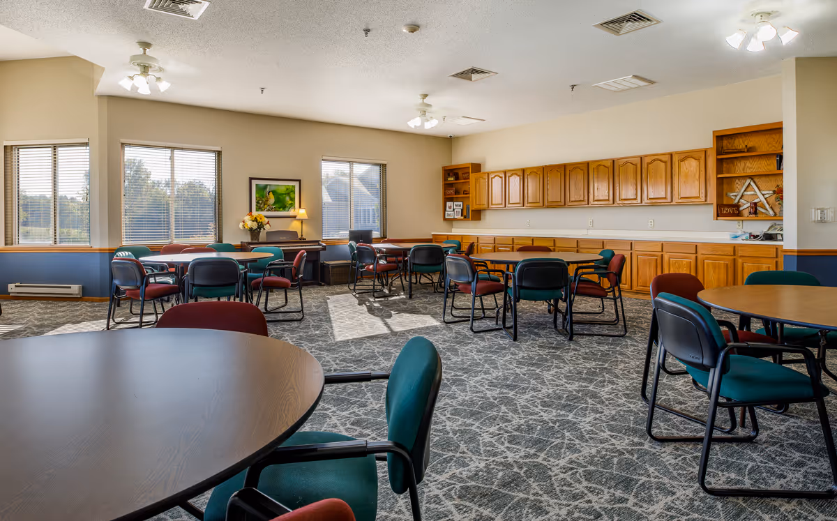 A spacious dining room in an assisted living facility with multiple round tables surrounded by green and red chairs. The room has large windows letting in natural light, a carpeted floor with a patterned design, wooden cabinets along one wall, and a piano with a lamp and flower arrangement near the windows.
