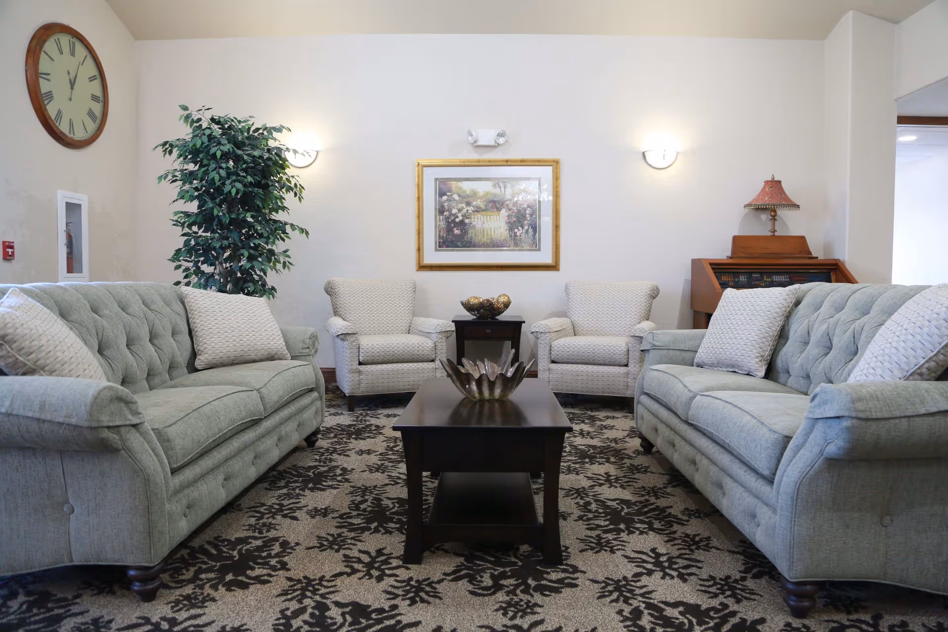 A cozy living room area with two light green tufted sofas facing each other, two beige armchairs, a dark wooden coffee table with a decorative bowl, a patterned carpet, a large wall clock, a framed floral painting, a potted plant, and a small wooden cabinet with a red lampshade.