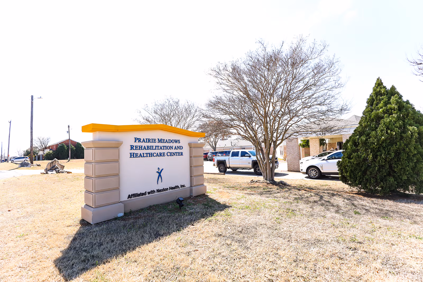 Outdoor front sign for Prairie Meadows Rehabilitation and Healthcare Center on a grassy lawn with the building and parked cars behind it.