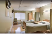 A hospital-style room with two beds separated by a curtain. Each bed has light green bedding and a wooden headboard. There is a chair near the window with blinds, a small cabinet, and a wall-mounted TV. The room has a clean, clinical appearance with beige walls and a tiled floor.