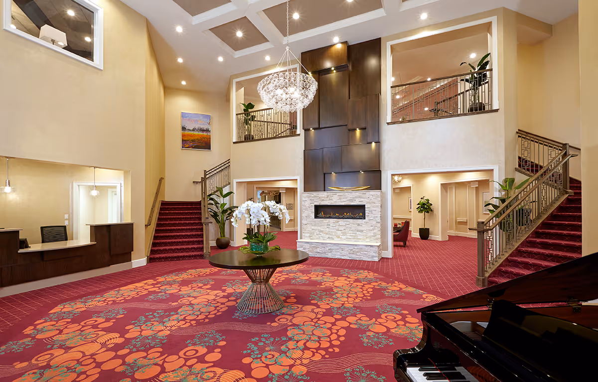 Spacious and elegant lobby area with a red patterned carpet, a round table with white orchids in the center, a modern fireplace with a dark wood panel backdrop, two staircases on either side leading to an upper level, a grand chandelier hanging from the ceiling, and a black grand piano in the foreground.