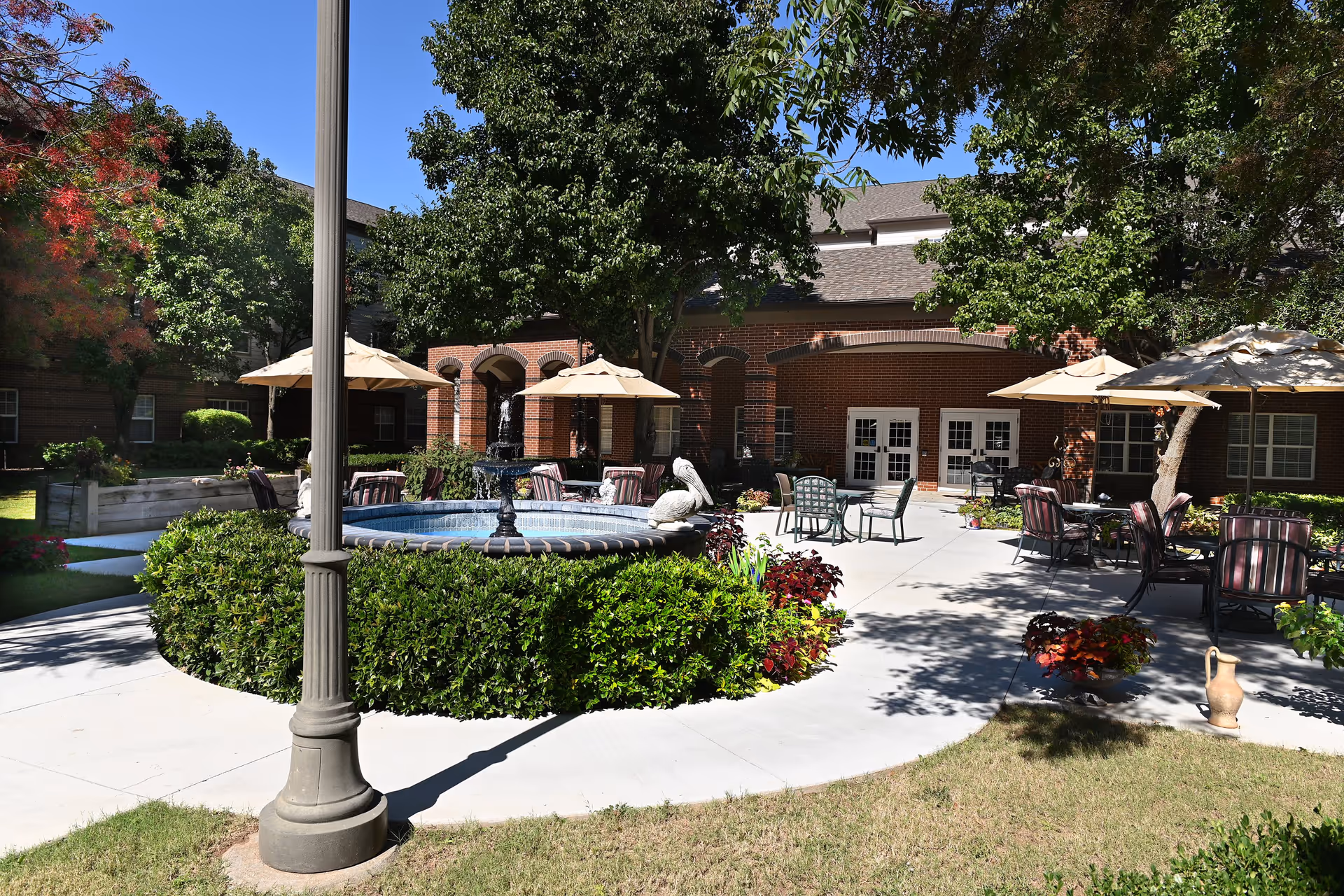 Outdoor courtyard area at Town Village Tulsa featuring a central water fountain surrounded by green bushes, multiple patio tables with umbrellas, chairs, and trees providing shade. The background shows a brick building with arched doorways and windows.