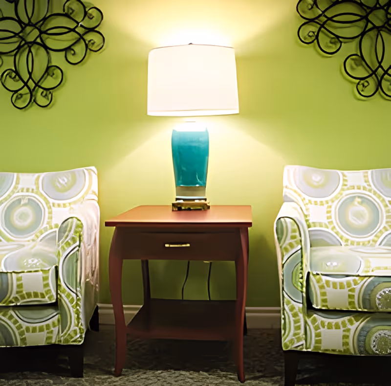 Two patterned armchairs with circular designs flank a wooden side table with a drawer. A blue ceramic table lamp with a white shade sits on the table, casting light on a green wall decorated with black wrought iron wall art on either side.