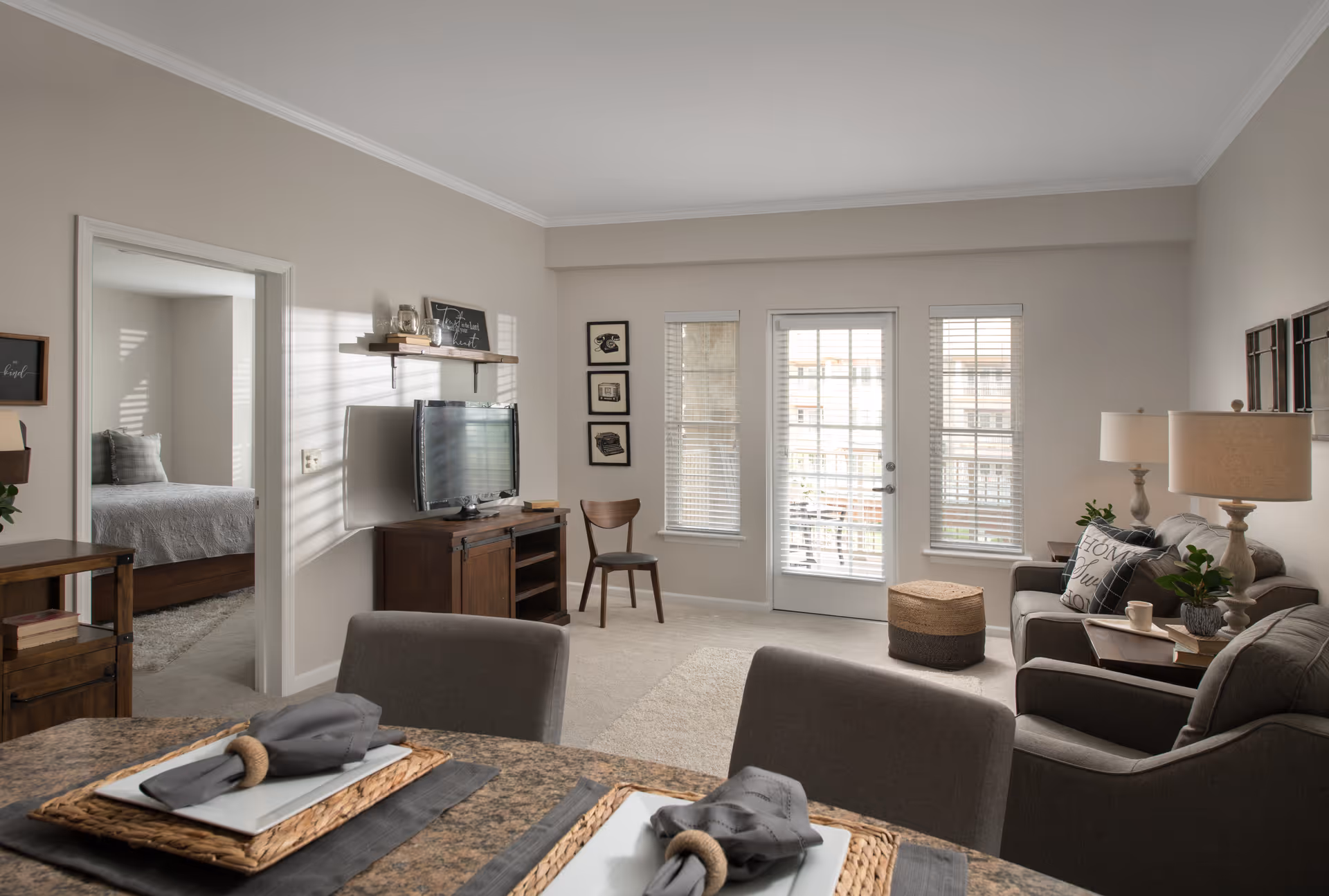 A cozy living room with neutral tones featuring a sofa, armchair, side tables with lamps, a TV on a wooden stand, and a dining table set with placemats and napkins. There is a door and two windows with blinds letting in natural light. A bedroom with a bed and pillows is visible through an open doorway.