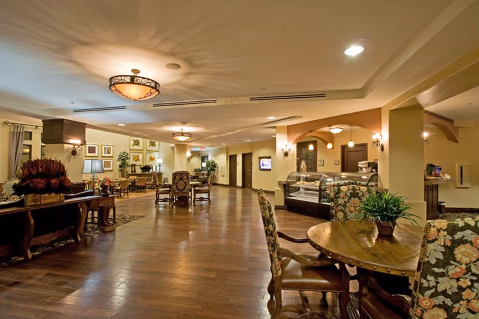 Spacious, warmly lit senior living common area with wooden floors, round tables and upholstered chairs, decorative lighting, and a display case.
