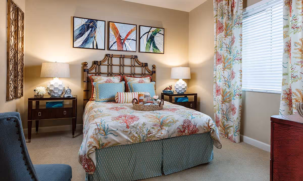 A cozy bedroom with a bed featuring a coral and seaweed patterned bedspread and matching curtains. The bed has a wooden headboard and is flanked by two nightstands, each with a white geometric lamp. Above the bed are three abstract paintings with colorful brush strokes. A window with white blinds is on the right side of the room, and a blue upholstered chair is partially visible on the left.