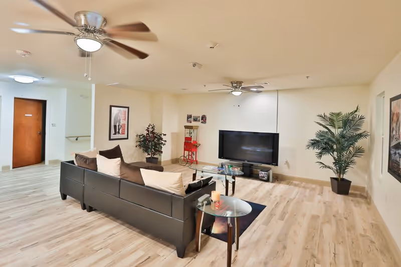 A spacious living room with light wood flooring, a black leather sectional sofa with beige and brown pillows, a glass coffee table, and a flat-screen TV mounted on the wall. The room has two ceiling fans with lights, a potted plant in the corner, framed artwork on the walls, and a small popcorn machine near the TV.