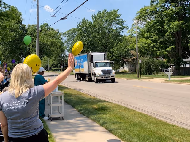People standing on a sidewalk waving yellow smiley face balloons at a white delivery truck driving down a tree-lined street on a sunny day.