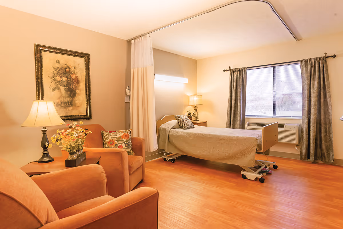 A warm, well-lit bedroom in a senior living facility featuring a hospital-style bed with a beige cover, a window with curtains and blinds, two comfortable armchairs with floral cushions, a side table with a lamp and a flower arrangement, and a framed painting of a floral bouquet on the wall.