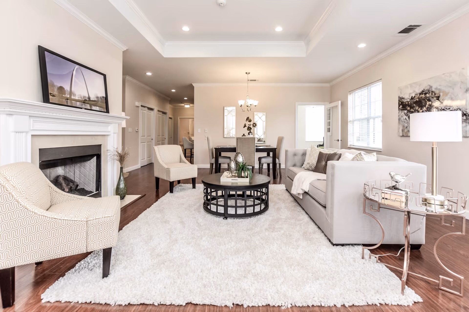 A bright and spacious living room with a light gray sofa, two beige armchairs, a round black coffee table on a fluffy white rug, a white fireplace with a framed picture above it, a side table with a lamp, and a dining area with a table and chairs in the background.