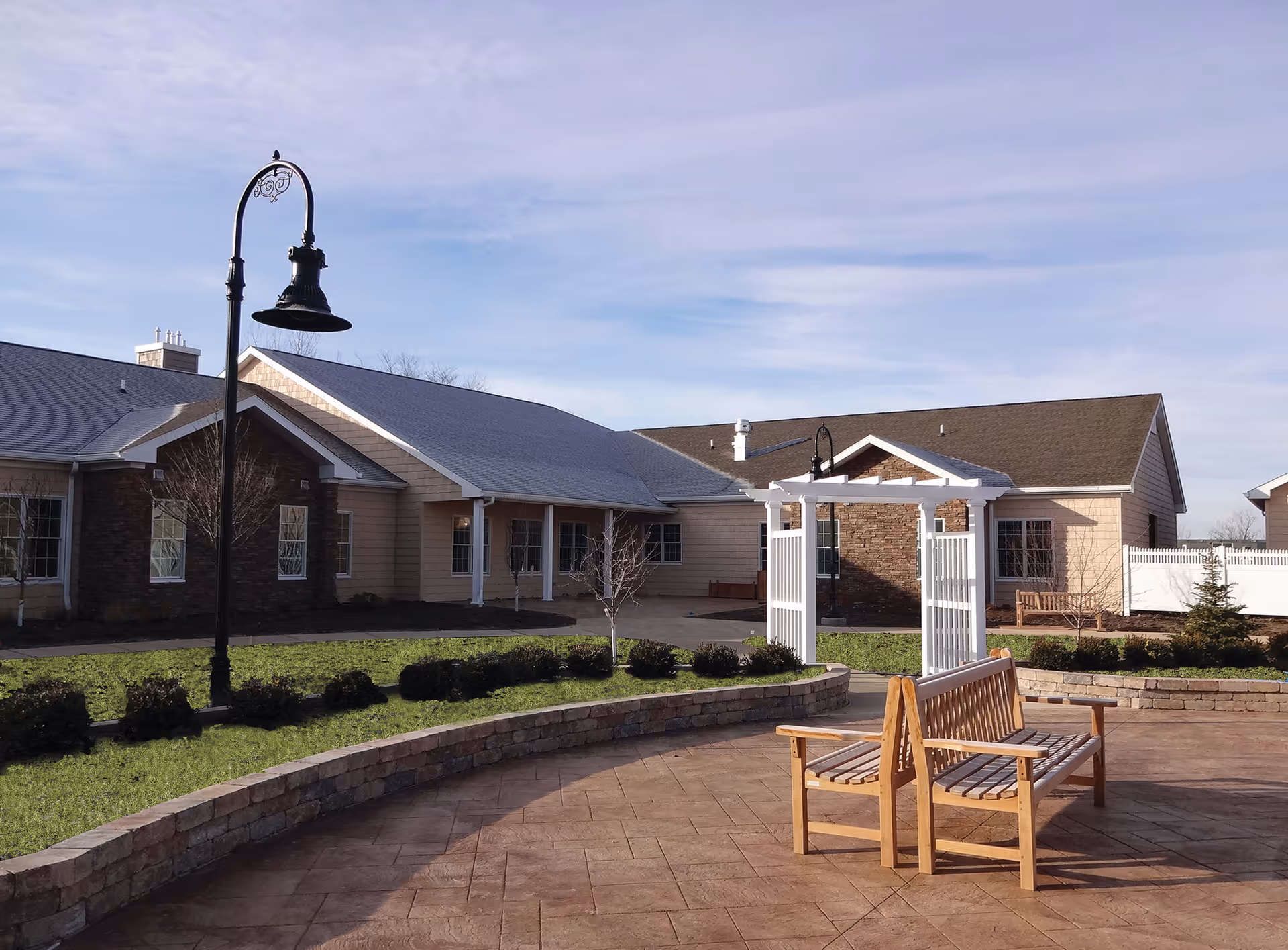 Outdoor courtyard with wooden benches, a lamp post, and a one-story senior living building under a blue sky.