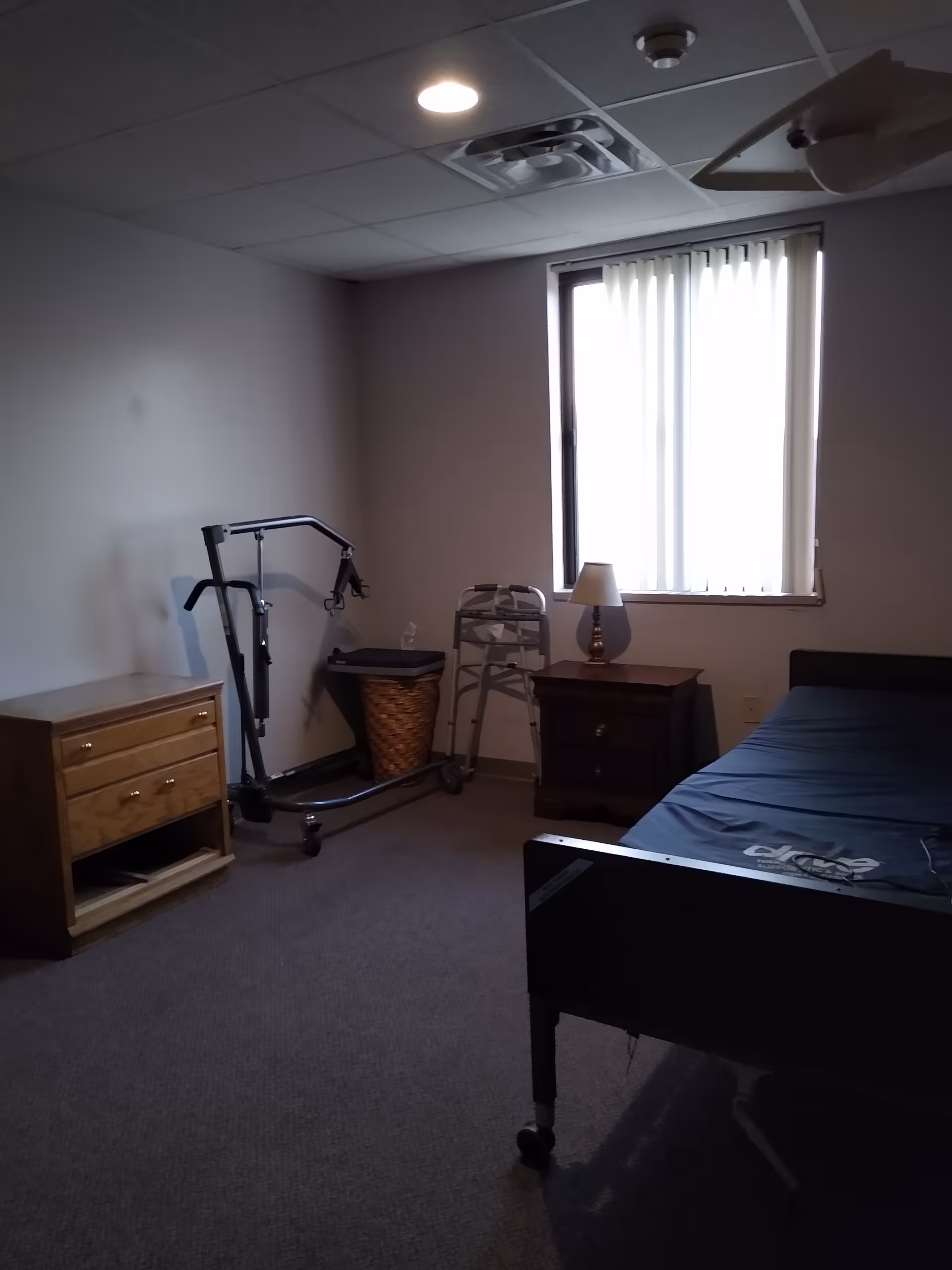 A simple bedroom with a hospital-style bed, a wooden nightstand with a lamp, a wooden dresser, a walker, a patient lift, and a window with vertical blinds letting in light.