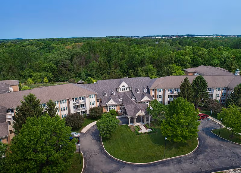 Aerial view of Baldwin House Senior Living Oakland, showing a large multi-wing building surrounded by trees and greenery with a circular driveway and well-maintained lawn in front.