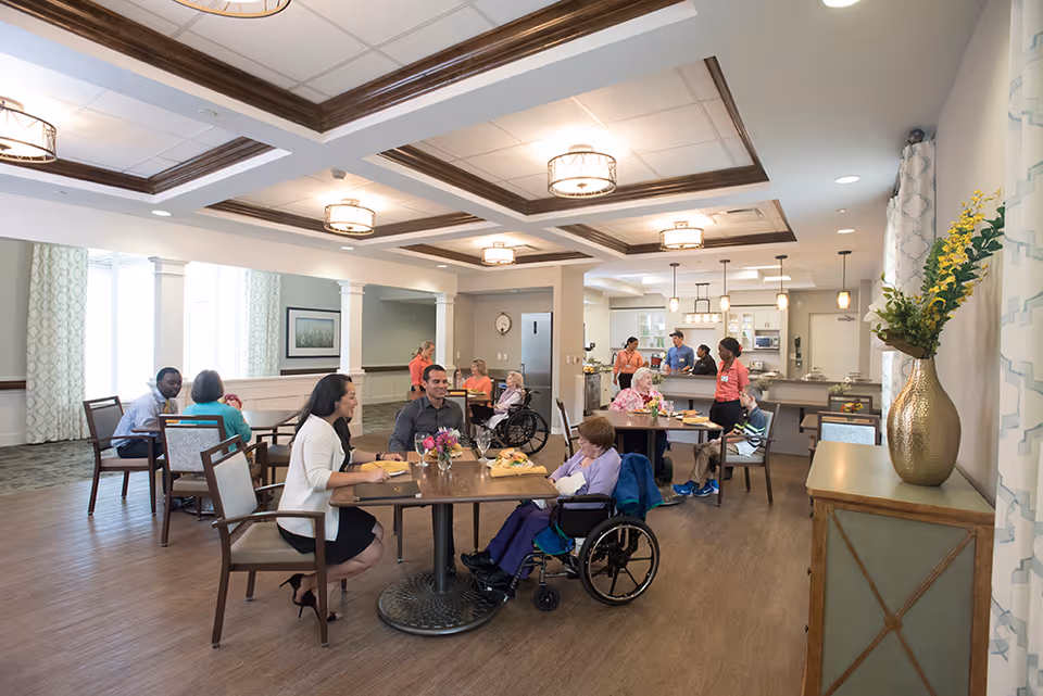 Spacious dining and communal area with residents and staff seated at tables near a serving counter and open kitchen.