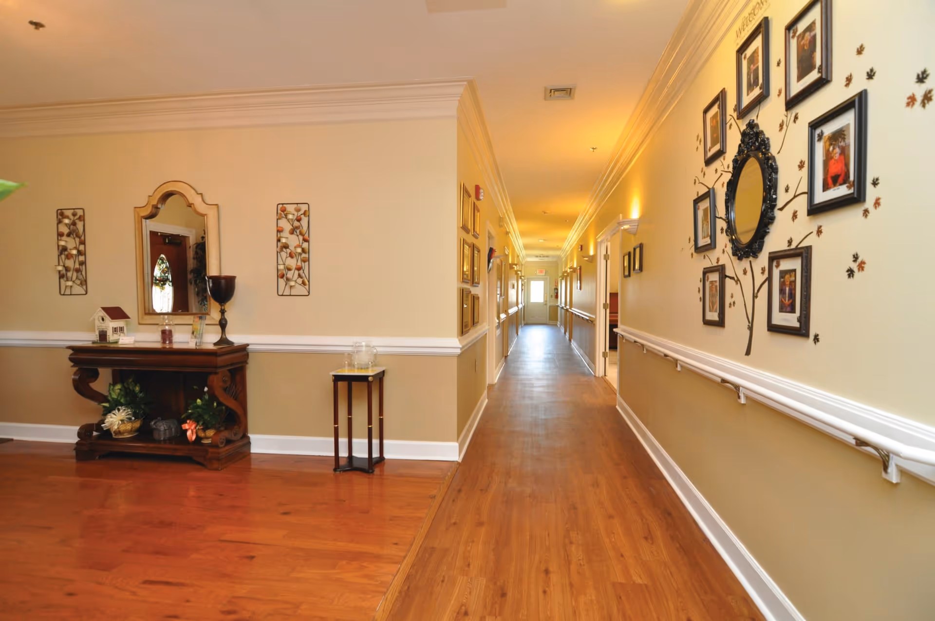 A well-lit hallway in Rose Wood Manor with wooden flooring and beige walls. On the left side, there is a wooden console table with decorative items and a mirror above it. On the right wall, there is a decorative arrangement of framed photos around a central mirror, with a tree and leaf design painted on the wall. Handrails run along both sides of the hallway, leading to a door at the far end.