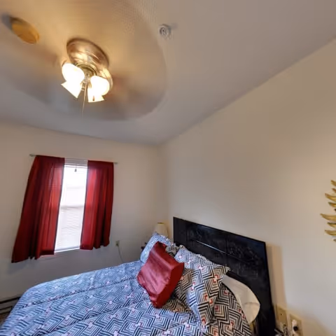 A bedroom with a bed covered in a geometric patterned bedspread and a red accent pillow. The bed has a dark headboard. There is a window with red curtains on the left wall and a ceiling fan with lights above.