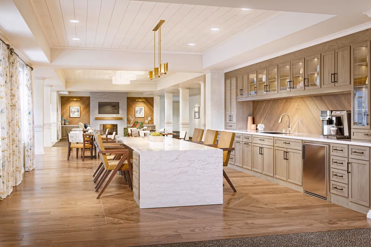 A spacious and modern communal kitchen and dining area with a large white marble island surrounded by wooden chairs. The kitchen features light wood cabinetry with glass doors, a built-in coffee machine, and a stainless steel mini fridge. The room has wooden flooring, recessed lighting, and large windows with floral curtains allowing natural light to fill the space. In the background, there is a cozy seating area with a fireplace and a wall-mounted TV.