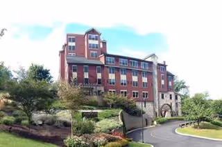 Exterior view of a multi-story senior living facility building with red brick and stone accents, surrounded by landscaped greenery and a curved driveway leading to the entrance.