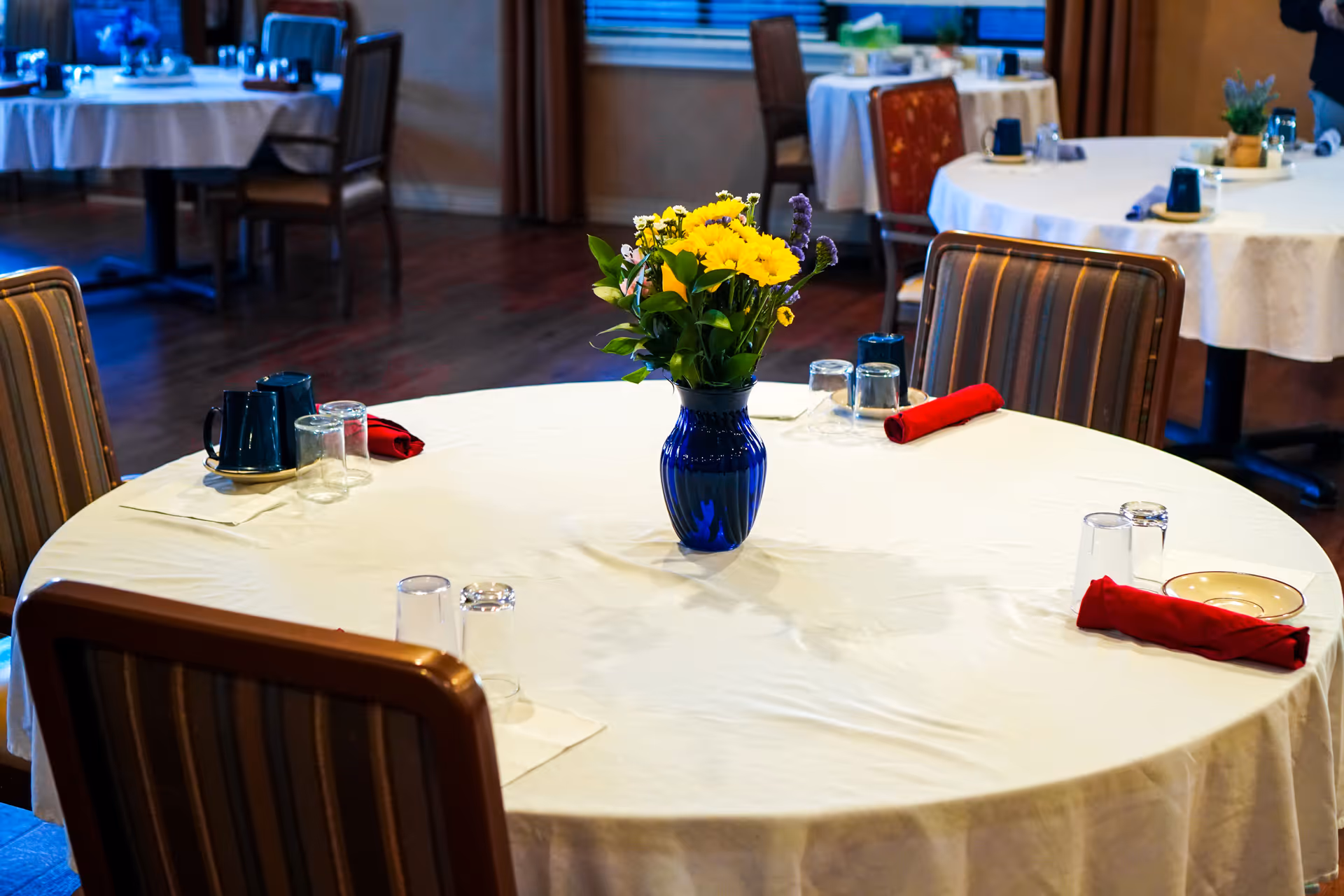 A dining room with round tables covered in white tablecloths, each set with glasses, red cloth napkins, and a blue vase with yellow flowers as a centerpiece. The room has wooden floors and striped cushioned chairs around the tables.