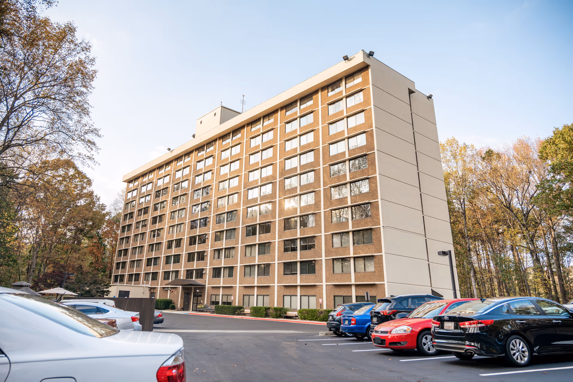 Exterior view of Smyrna Towers Senior Apartments, a multi-story residential building with numerous windows. Several cars are parked in the parking lot in front of the building. Trees with autumn foliage surround the area under a clear sky.