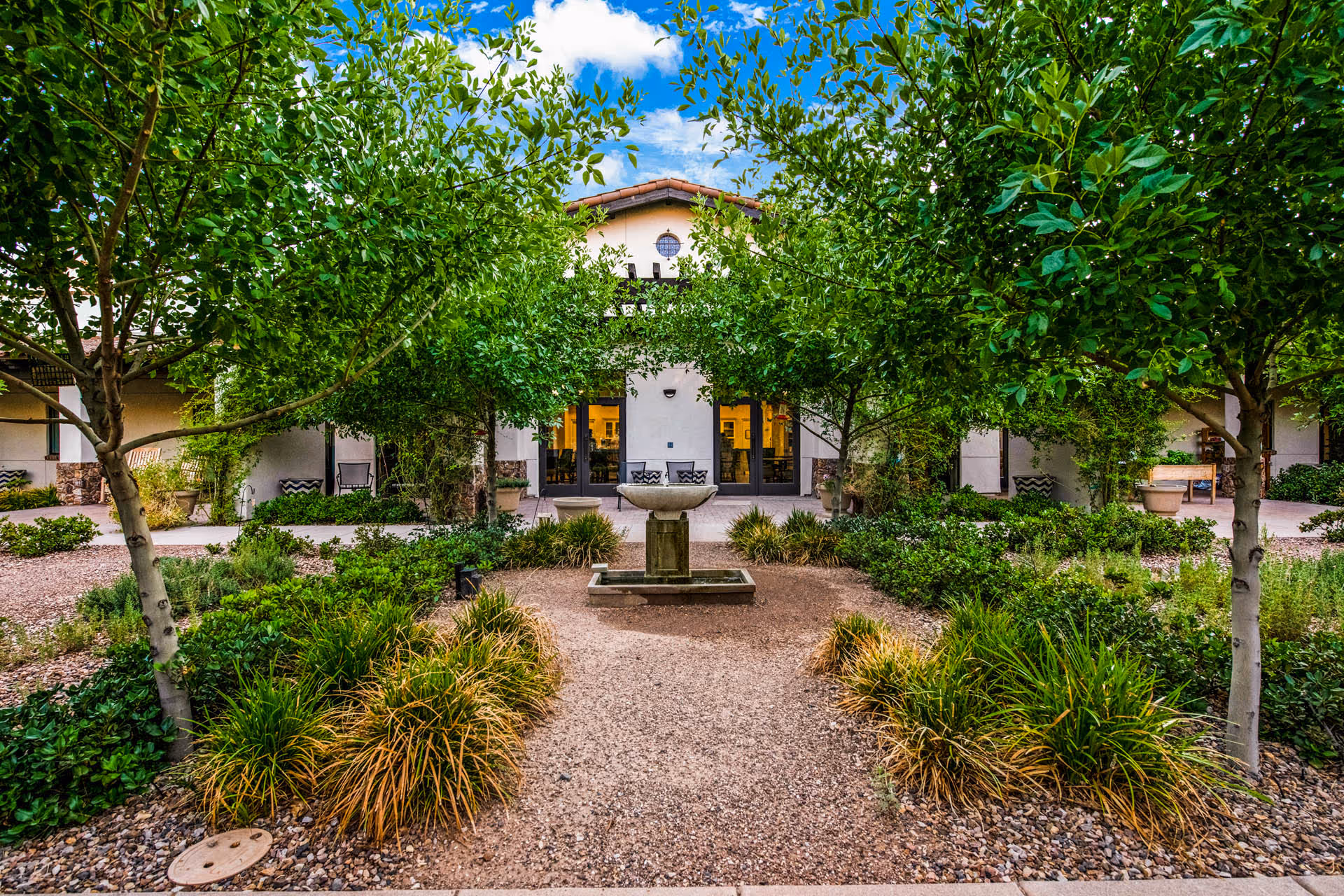 A landscaped outdoor garden area with a gravel pathway leading to a central stone fountain. The garden is surrounded by green bushes and trees, with a building featuring large glass doors and windows in the background under a partly cloudy blue sky.