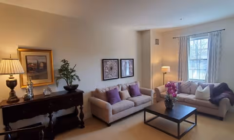 A cozy living room with two beige sofas adorned with purple and white pillows, a dark wooden coffee table with a flower arrangement, a side table with a lamp and decorative items, framed artwork on the wall, and a window with light curtains letting in natural light.