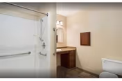 A clean and simple bathroom featuring a white shower with grab bars, a vanity with a sink and mirror, and a toilet. The walls are painted beige and the floor has a dark tile finish.