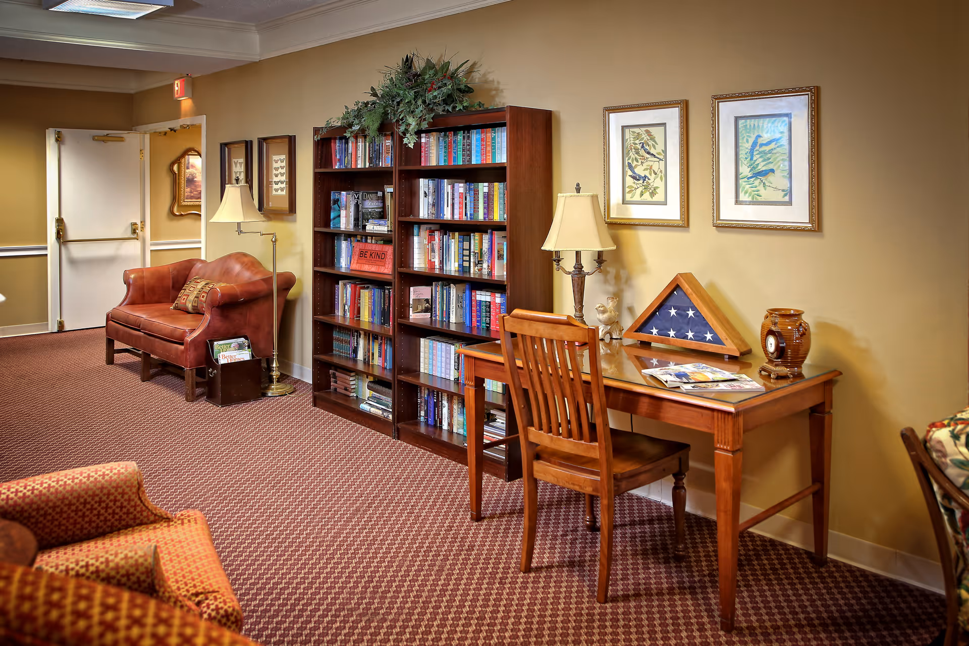 Cozy common room with bookshelves, a wooden desk and chair, lamps, armchairs, and a leather loveseat.