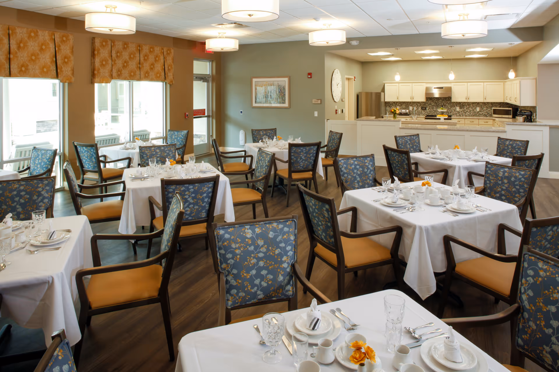 A bright dining room with multiple tables covered in white tablecloths, each set with plates, glasses, silverware, and napkins. The chairs have wooden frames with blue floral upholstery and tan seats. Large windows with patterned valances let in natural light. In the background, there is a kitchen area with white cabinets, a refrigerator, and a stove. The room has a warm and inviting atmosphere.