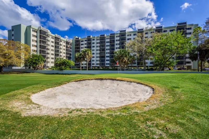 A large multi-story senior living facility building with balconies, surrounded by trees and greenery. In the foreground, there is a sand bunker and a putting green, indicating a golf course area in front of the building under a partly cloudy blue sky.