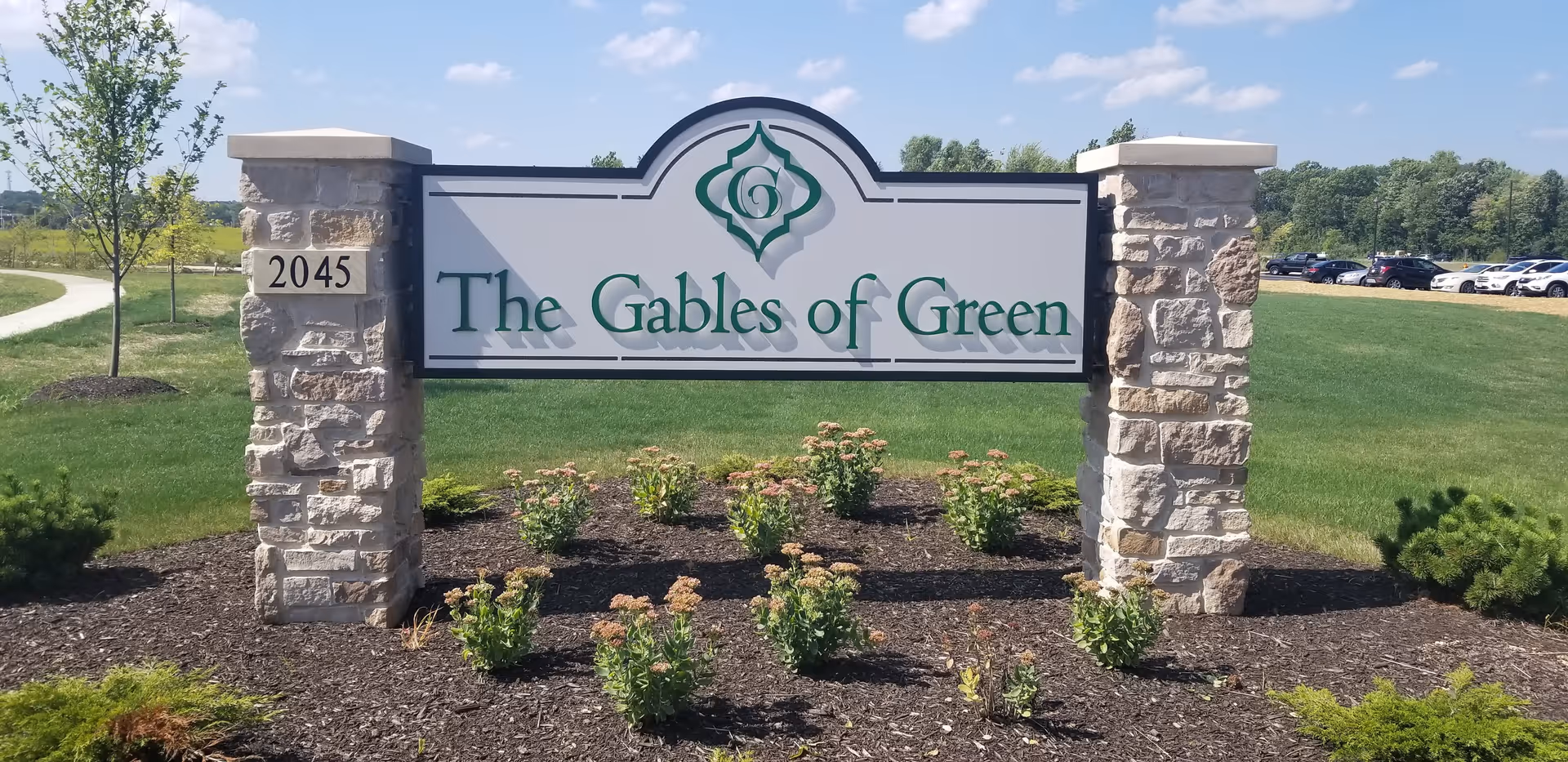 Stone pillars supporting a sign that reads 'The Gables of Green' with a landscaped area of small bushes and mulch in front, a grassy field, trees, and a parking lot with cars in the background under a blue sky with some clouds.