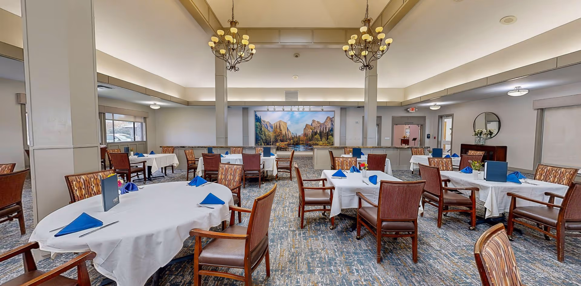 A spacious dining room with multiple tables covered in white tablecloths and set with blue folded napkins. The room features wooden chairs with patterned upholstery, two large chandeliers hanging from the ceiling, and a large scenic mural on the far wall depicting a mountain landscape. The room has large windows and neutral-colored walls.