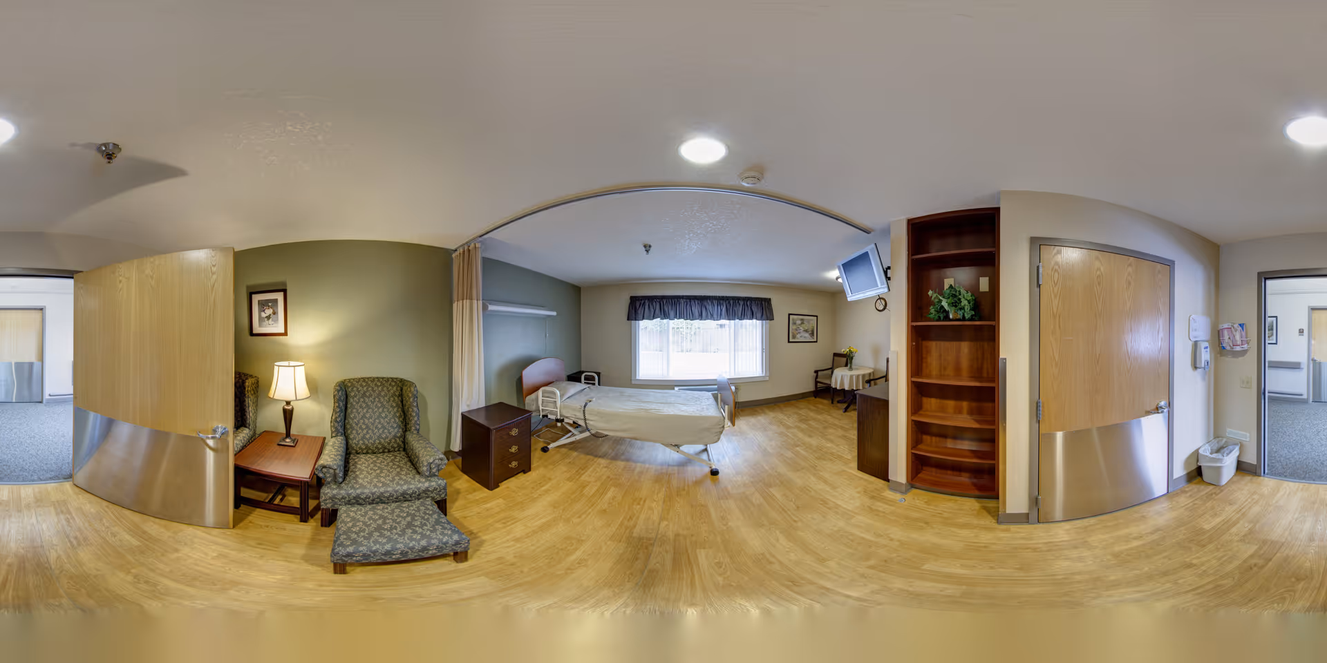 A well-lit patient room in a senior living facility featuring a hospital bed near a window with a dark valance, a green patterned armchair with matching ottoman beside a wooden side table with a lamp, a wall-mounted TV, wooden shelving, and two wooden doors leading to adjacent rooms or hallways. The room has light wood flooring and light-colored walls with some green accent walls.