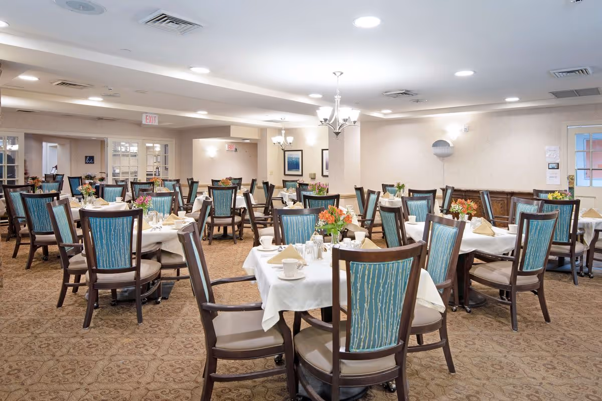 Spacious dining room with multiple round tables set with white linens, floral centerpieces, and teal-upholstered chairs.