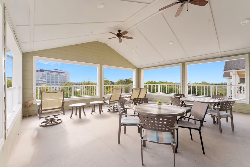 Covered outdoor patio area with multiple round tables and chairs, ceiling fans, and large open windows offering views of trees and a distant building under a clear blue sky.
