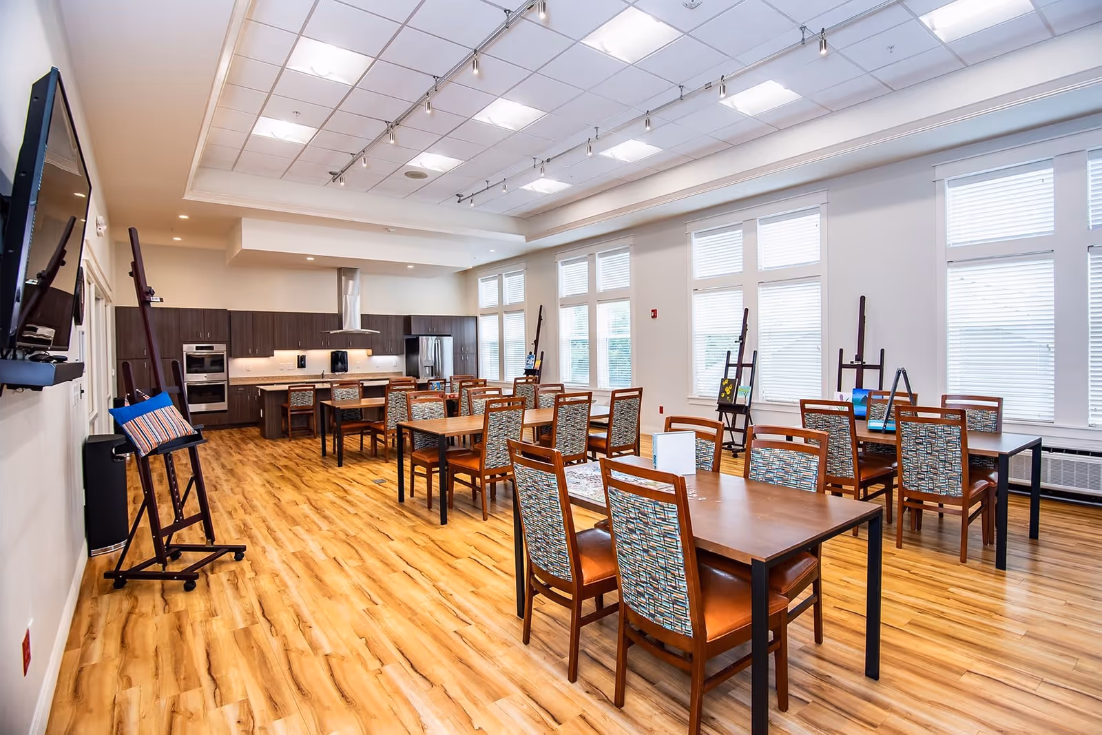 A bright and spacious room with wooden flooring featuring multiple tables and chairs arranged for group seating. The room has large windows with white blinds, a modern kitchen area with dark cabinets, stainless steel appliances, and a range hood. Several easels with paintings are positioned near the windows, and a large flat-screen TV is mounted on the wall.