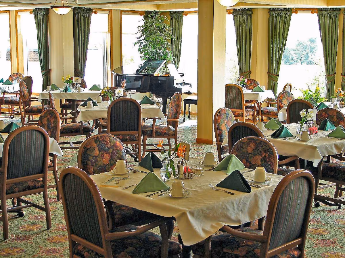A bright dining room with multiple tables covered in white tablecloths, each set with green folded napkins, cups, and silverware. The chairs have floral and striped upholstery. Large windows with green curtains let in natural light, and a black grand piano is visible near the windows.