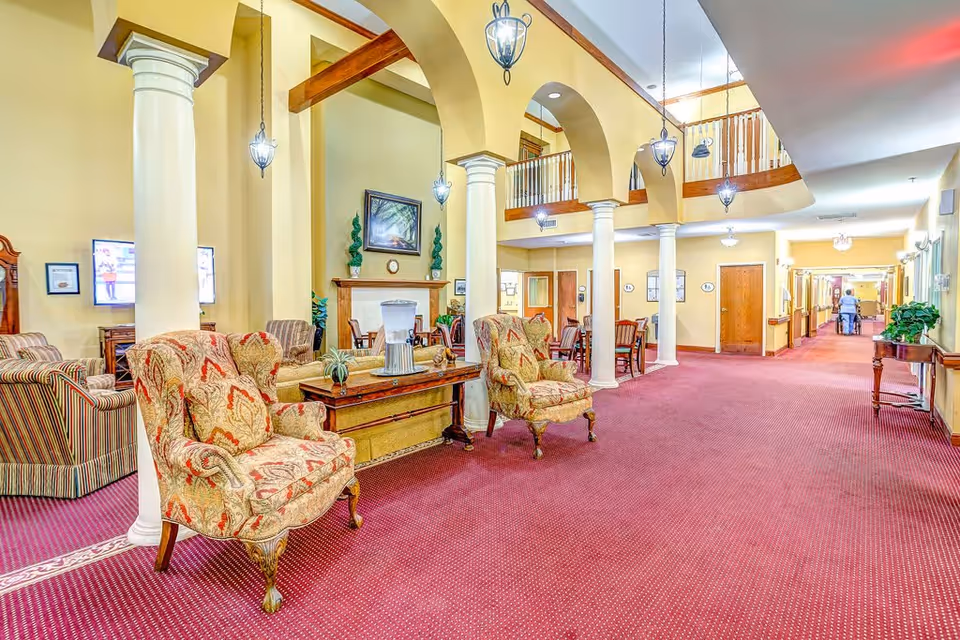 A spacious senior living facility common area with red carpet flooring, beige walls, and white columns. The area features patterned armchairs, a wooden table with a water dispenser, and a fireplace with decorative plants and a painting above it. There are hanging lantern-style lights and a balcony railing visible on the upper level. In the background, a hallway extends with doors and a person walking with a walker.