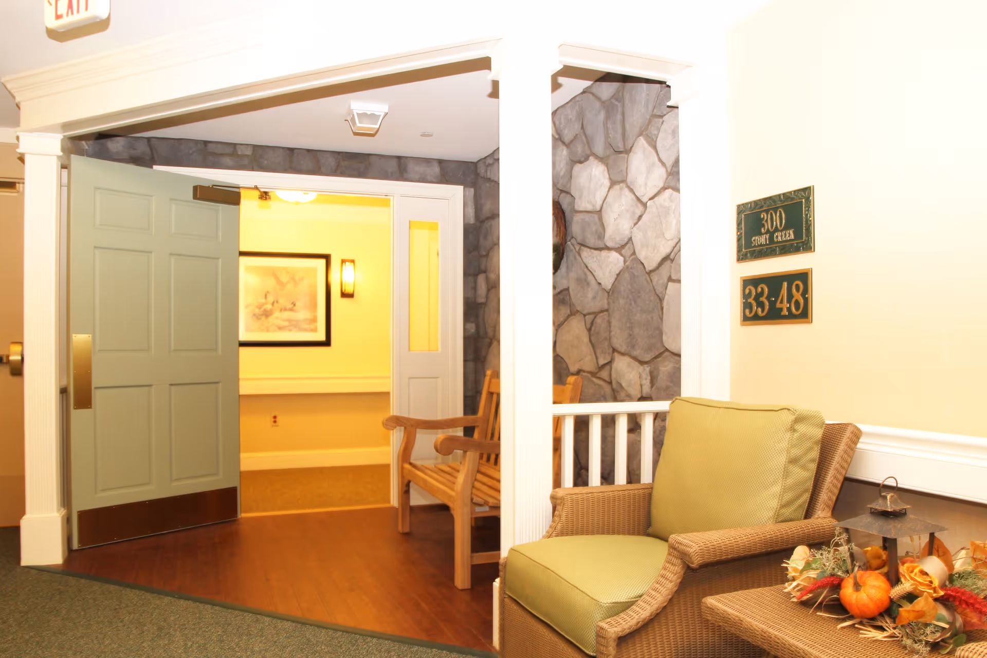 A cozy interior hallway area with a green cushioned wicker chair and a small table decorated with autumn-themed items including small pumpkins and flowers. There is a wooden bench in the background near a stone wall, and an open green door leading to a warmly lit room with a framed picture on the wall. Signs on the wall indicate room numbers 33-48 and 300 Stony Creek. An exit sign is visible above the doorway.