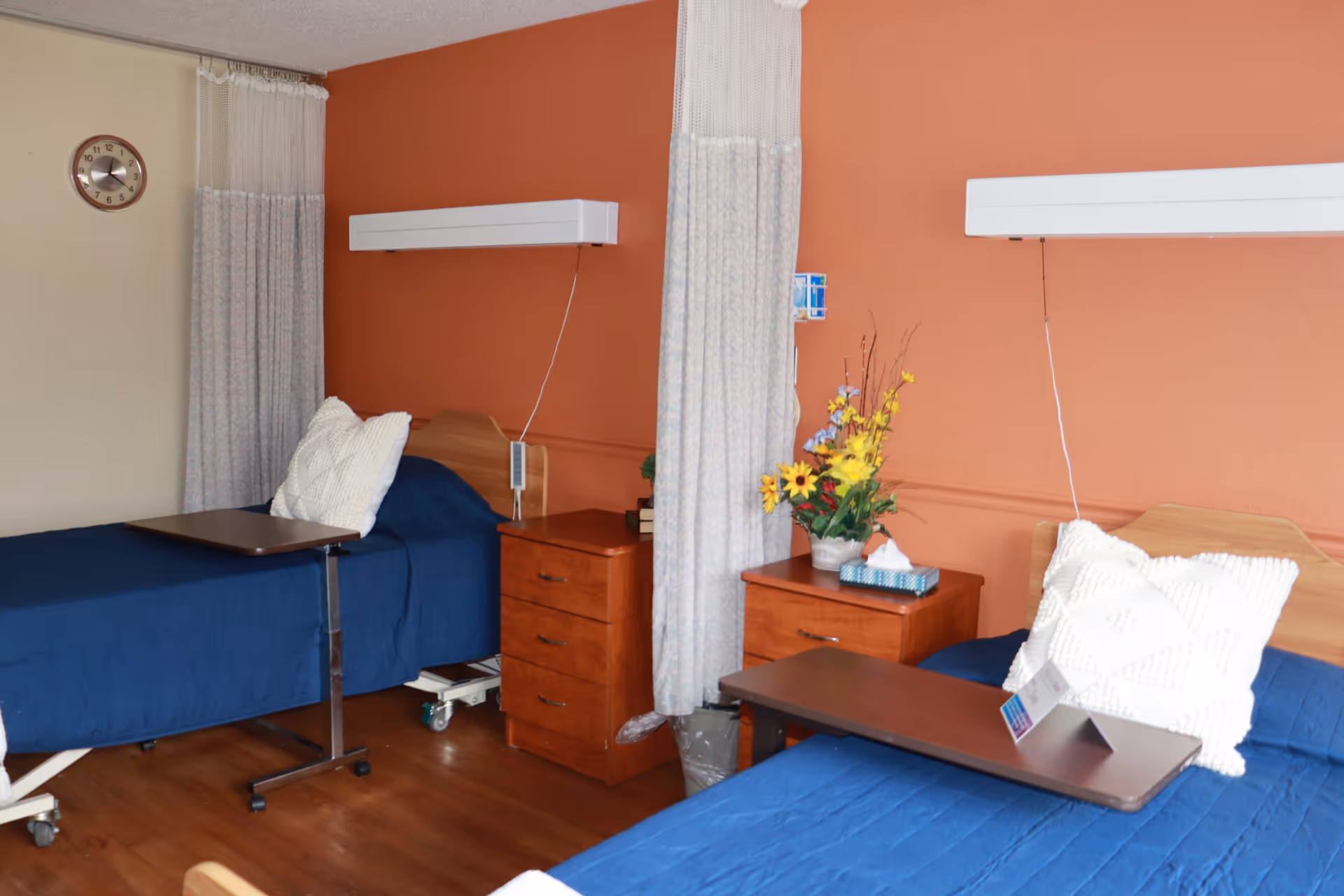 A room with two hospital-style beds, each with a blue bedspread and a white textured pillow. Between the beds are two wooden nightstands, one with a vase of yellow and purple flowers and a box of tissues. There are privacy curtains hanging between the beds and a clock on the wall. The walls are painted in a warm orange and beige color, and the floor is wooden.
